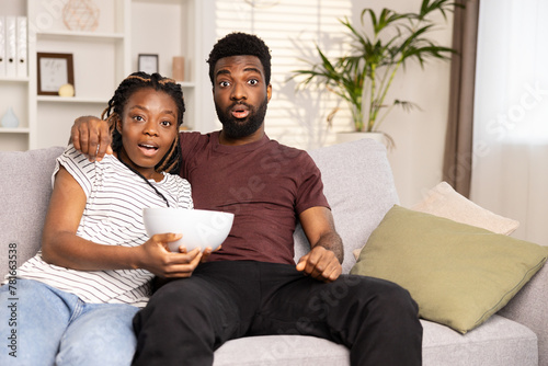 Surprised Afro Couple Watching TV On Couch With Snacks. Home Entertainment, Shock, Excitement, Leisure Time Concepts. Captured In Cozy Living Room. Perfect For Ads And Social Media Content.