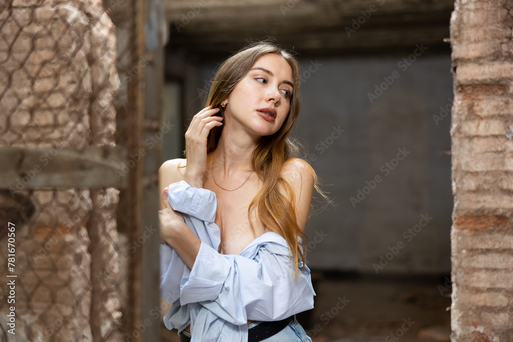 Tempting young brown-haired woman in blue male shirt pulled down from her shoulders and jeans standing near old rusty wire fencing in dilapidated abandoned house