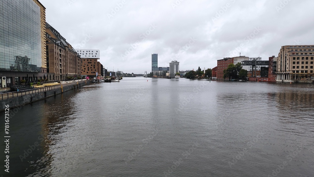 Cloudy cityscape with riverfront buildings and calm water under an overcast sky.