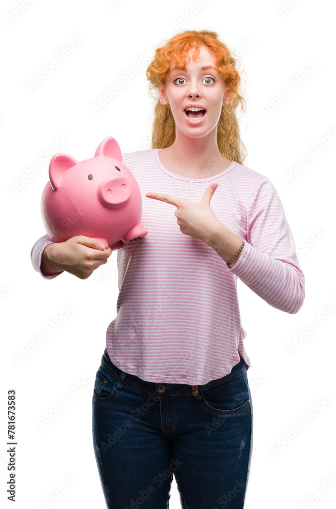 Young redhead woman holding piggy bank very happy pointing with hand and finger Young redhead woman holding piggy bank very happy pointing with hand and finger