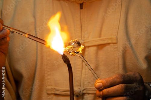 A man is holding a glass object that is being heated by a flame. Concept of craftsmanship and skill, as the man carefully handles the hot glass. The use of fire in the process adds a dramatic photo