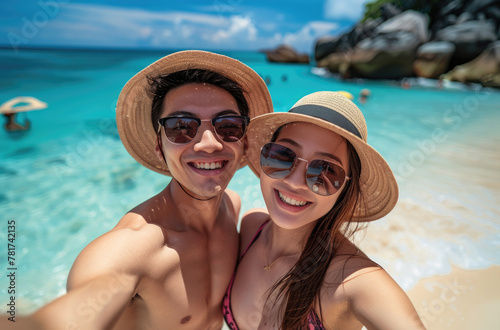 A young Asian couple taking selfie at tropical beach on vacation, wearing sun hats and sunglasses while having fun together in summer holiday trip © Kien