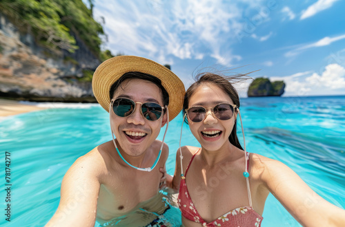 A young Asian couple taking selfie at tropical beach on vacation, wearing sun hats and sunglasses while having fun together in summer holiday trip © Kien