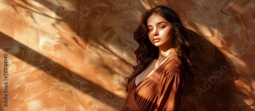 A woman wearing a brown dress is standing gracefully in front of a textured wall, showcasing elegance and poise