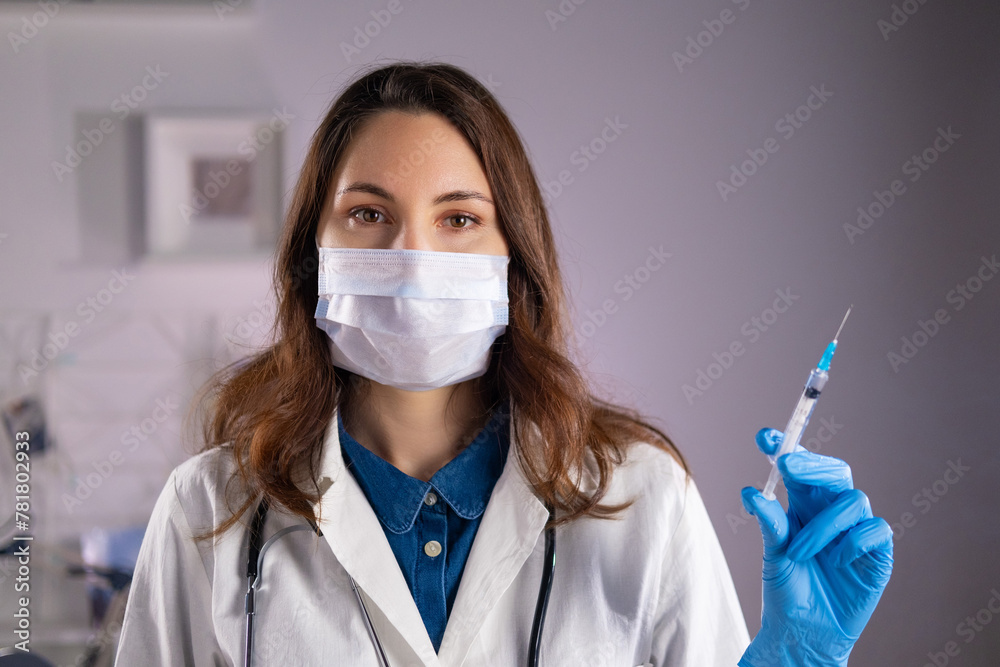 A young adult female doctor in a medical mask and white coat holds a syringe in her hands and looks at the camera A young adult female doctor in a medical mask and white coat holds a syringe in her hands and looks at the camera