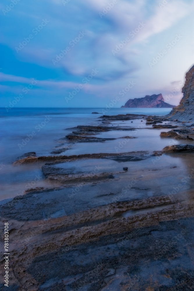 Sunset at Cala Baladrar beach with the Peñon de Ifach in the background and rocks in the foreground. Sea of silk. Long exposure