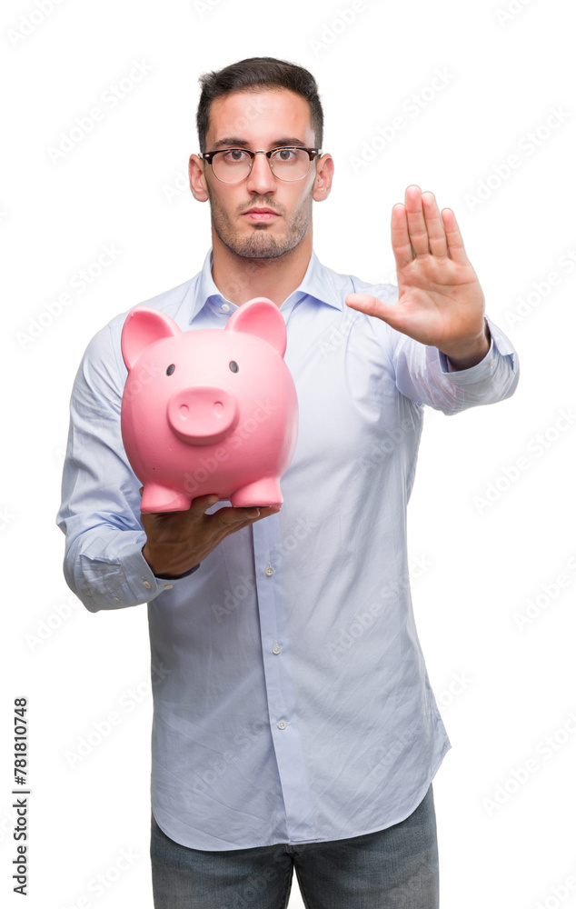 Handsome young man holding a piggy bank with open hand doing stop sign with serious and confident expression, defense gesture Handsome young man holding a piggy bank with open hand doing stop sign with serious and confident expression, defense gesture