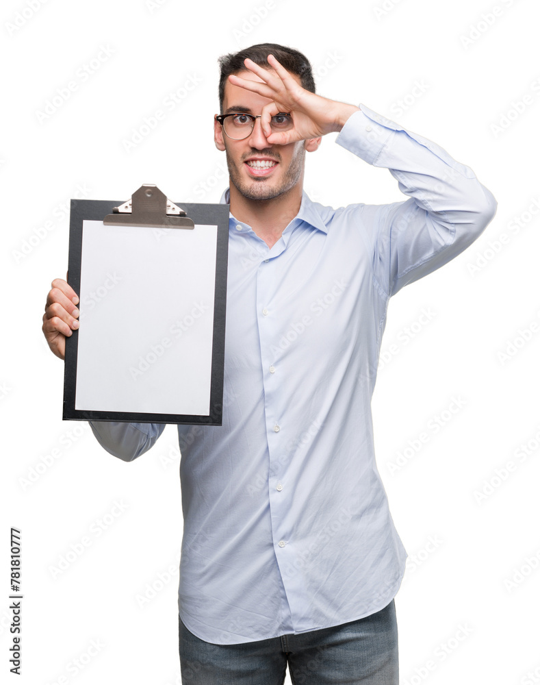 Handsome young business man showing a clipboard with happy face smiling doing ok sign with hand on eye looking through fingers Handsome young business man showing a clipboard with happy face smiling doing ok sign with hand on eye looking through fingers