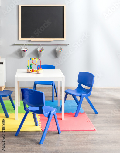 Bright and Colorful Kindergarten Classroom with a Chalkboard for Drawing, Blue Child-sized Plastic Chairs, White Activity Table, Educational Toys, Foam Floor Mat, Creating a Fun Learning Environment.