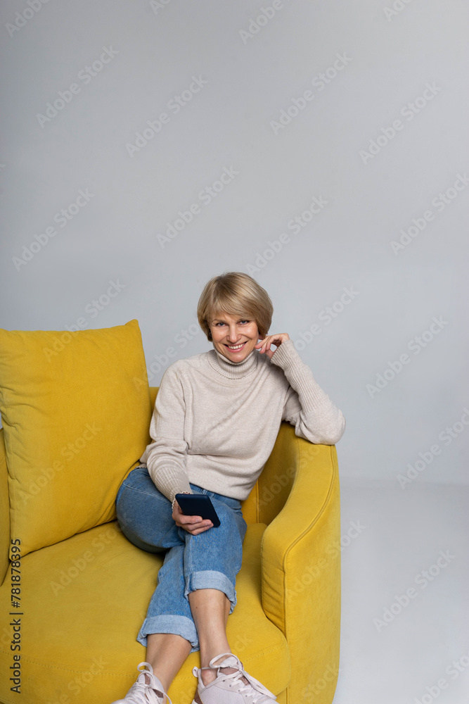 An adult lady sitting on the couch holding phone and smiling to all An adult lady sitting on the couch holding phone and smiling to all