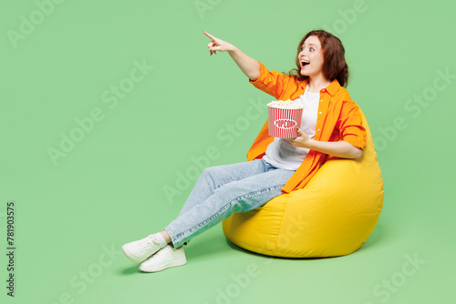 Full body young ginger woman she wear orange shirt white t-shirt casual clothes sit in bag chair eat pop corn watch movie film point finger aside isolated on plain green background. Lifestyle concept.