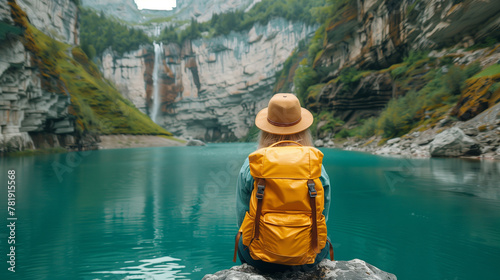 woman in hiking with yellow backpack and hat sits on the edge of an emerald lake looking at waterfalls