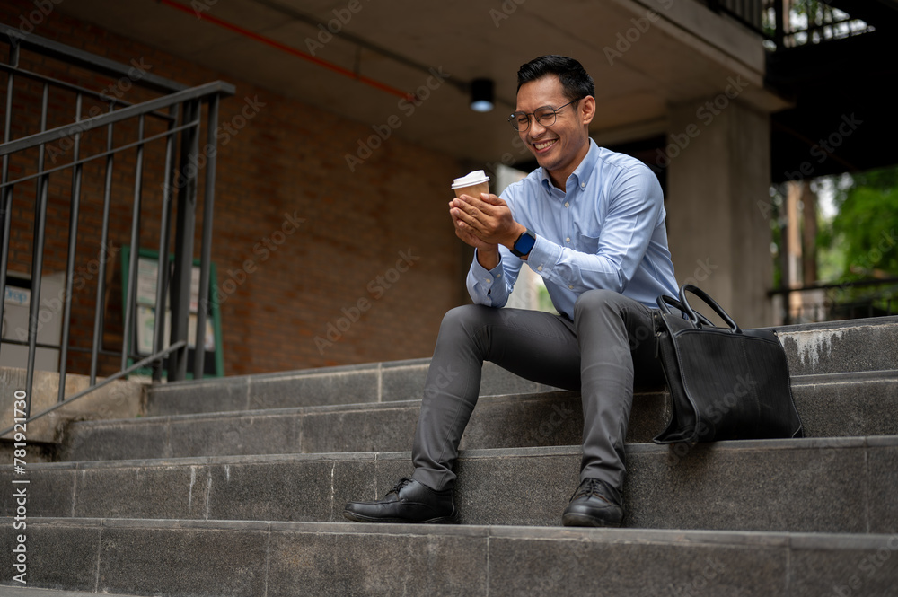 A handsome millennial Asian businessman enjoying his coffee on the stairs in front of the building. A handsome millennial Asian businessman enjoying his coffee on the stairs in front of the building.