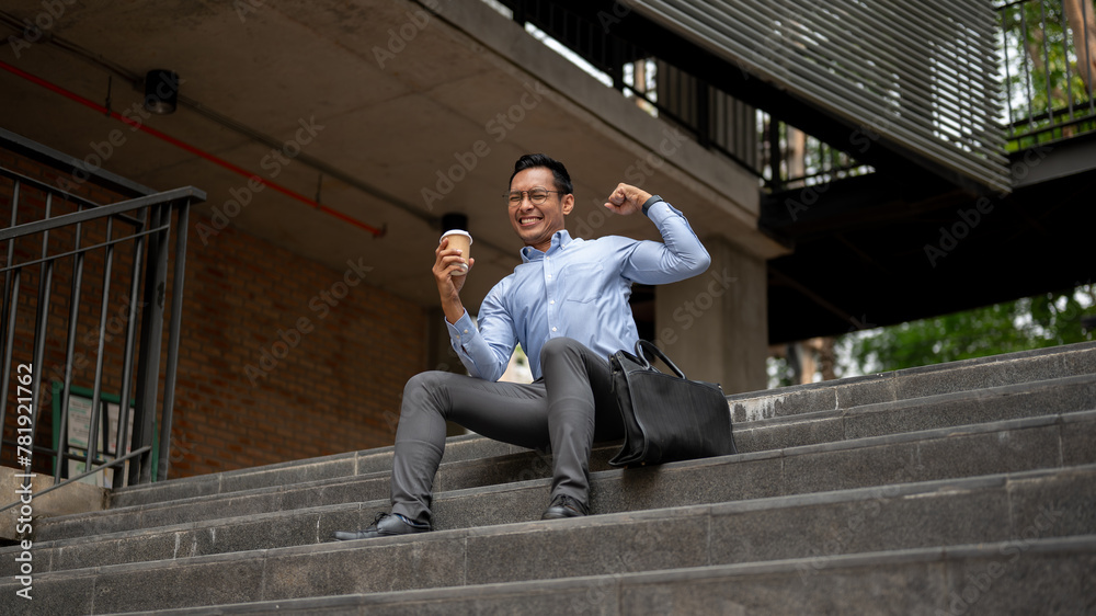 A happy Asian businessman sits on steps, holding a coffee cup and cheering with a raised fist. A happy Asian businessman sits on steps, holding a coffee cup and cheering with a raised fist.