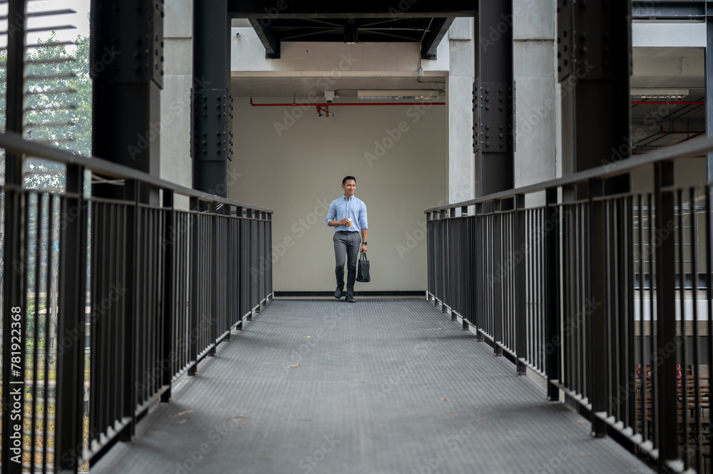 A handsome Asian businessman walking on a building corridor, holding a coffee cup and a briefcase.