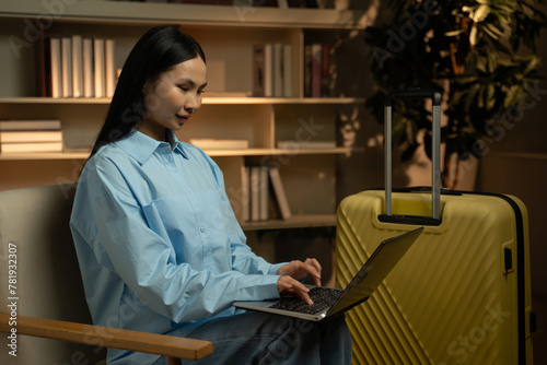 A young Asian woman is engrossed in using her laptop, sitting comfortably by a bright yellow suitcase, possibly indicating preparations for an upcoming trip or a return from travel, highlighted by the