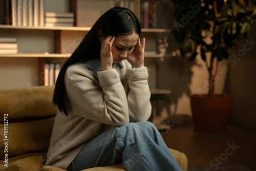 A young Asian woman is seated on a mustard-yellow armchair, clutching her head in apparent distress, indicating symptoms of a painful headache. She is alone in a warmly lit room that suggests a