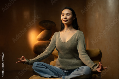A calm Asian woman is captured engaging in a peaceful mindfulness meditation session indoors with soft lighting that enhances the tranquil atmosphere, creating a synergetic blend of serenity and