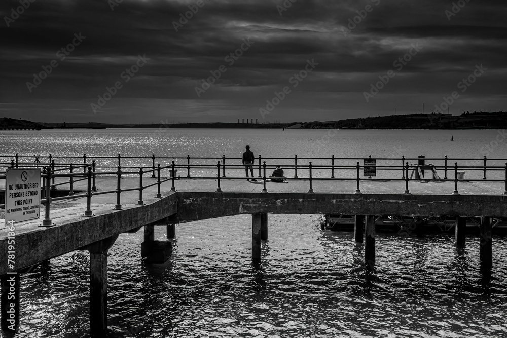 Fototapeta premium Scenic grayscale shot of people standing on a wooden pier and fishing in the tranquil sea