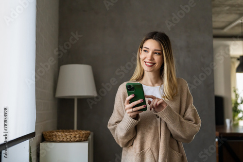 Young woman wearing sweater using smartphone at home, communication and social network concept, © NDABCREATIVITY