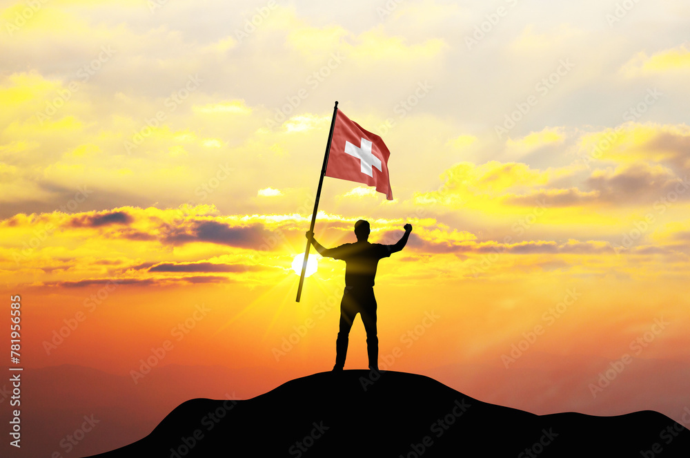 Switzerland flag being waved by a man celebrating success at the top of a mountain against sunset or sunrise. Switzerland flag for Independence Day.