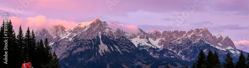 Panoramic shot of a mountain range covered with snow with pine forests in valleys at sunrise