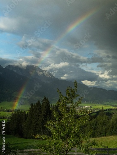 Vertical shot of a rainbow on a green field surrounded by high mountains