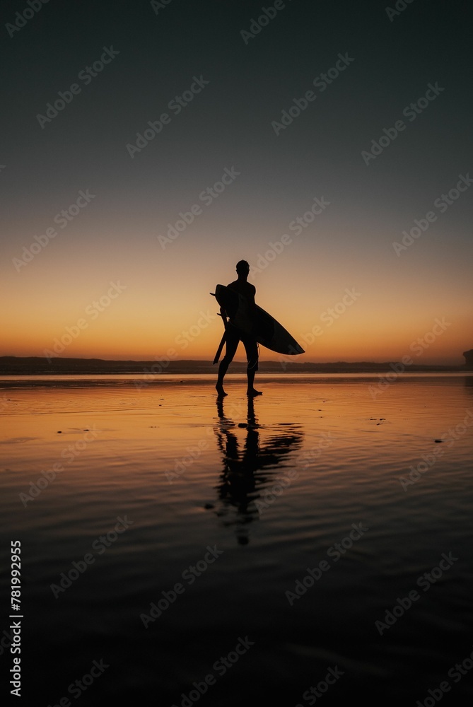 Vertical shot of a person with a surfing board walking on the beach at sunset perfect for wallpapers Vertical shot of a person with a surfing board walking on the beach at sunset perfect for wallpapers