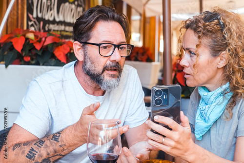 Modern adult couple look on phone contents sitting at the restaurant table with worried expression. Woman show cell to a man. Unhappy tourist writing a bad review after lunch. Problems notification