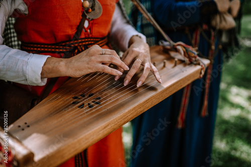 Canvas Print Valmiera, Latvia - July 14, 2023 - Hands playing a kokle, a traditional Latvian stringed musical instrument, with musicians in folk costumes blurred in the background