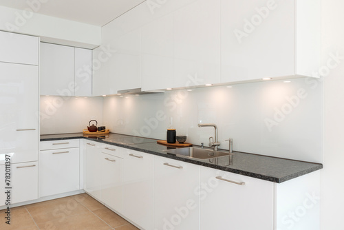 Glossy white minimalist modern kitchen with a black marble countertop. Spotlights illuminate the countertop, above which are two trays, one with a teapot and one with a vase
