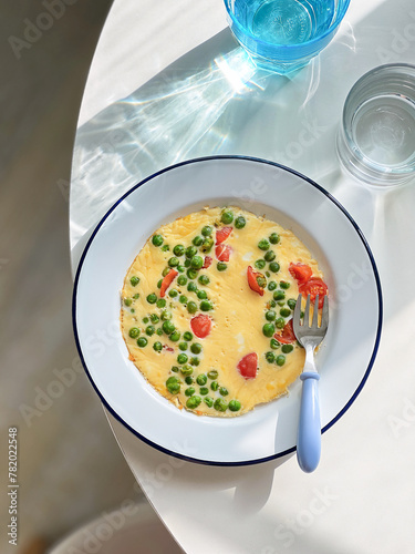 Breakfast omelette with green peas, cherry tomatoes on a plate. Top view. Blue glass of water. Natural light