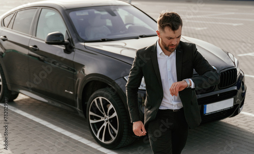 Checks the time. Businessman in suit is near his black car outdoors © standret