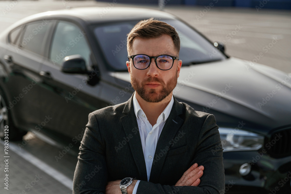 In glasses, front view. Businessman in suit is near his black car outdoors In glasses, front view. Businessman in suit is near his black car outdoors