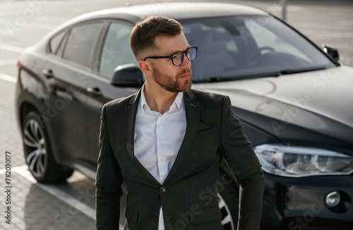 In glasses, front view. Businessman in suit is near his black car outdoors © standret
