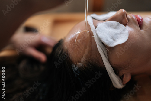 Closeup face of tranquil young woman relaxes in private room while receiving stress-relieving Shirodhara oil treatment at Ayurvedic retreat. Concept of luxury spa procedure. photo