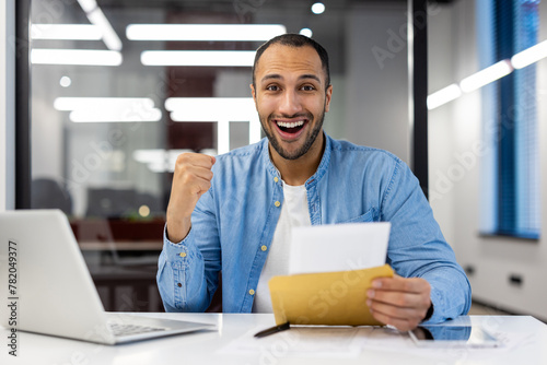 Portrait of a sad young Muslim man sitting in the office at the desk, holding an open envelope with a letter in his hands and looking happy at the camera, showing a victory gesture with his hand