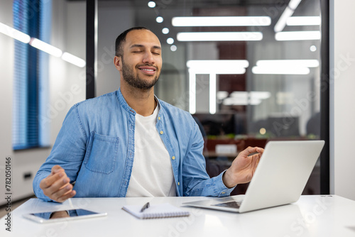 Smiling and relaxed young hispanic man sitting at desk in office in lotus position and meditating with closed eyes