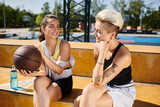 Two athletic women sitting side by side, holding a basketball, enjoying a summer day outdoors.