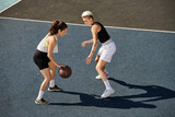 Two athletic young women playing basketball on an outdoor court in the summer.