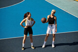 Two athletic young women, friends, stand confidently on top of a basketball court, enjoying a summer day.