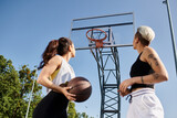 Two athletic young women stand together, holding a basketball, ready to play a game outdoors on a sunny day.