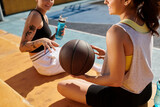 Two young women, athletic friends, sitting side by side holding a basketball outdoors on a sunny day.