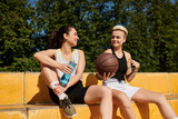 Two young women sit closely, holding a basketball, enjoying a game outdoors on a sunny summer day.
