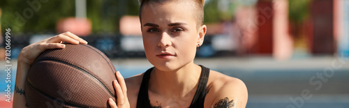 A young woman with short hair and tattoos confidently holds a basketball in her right hand, ready to showcase her athletic skills outdoors.