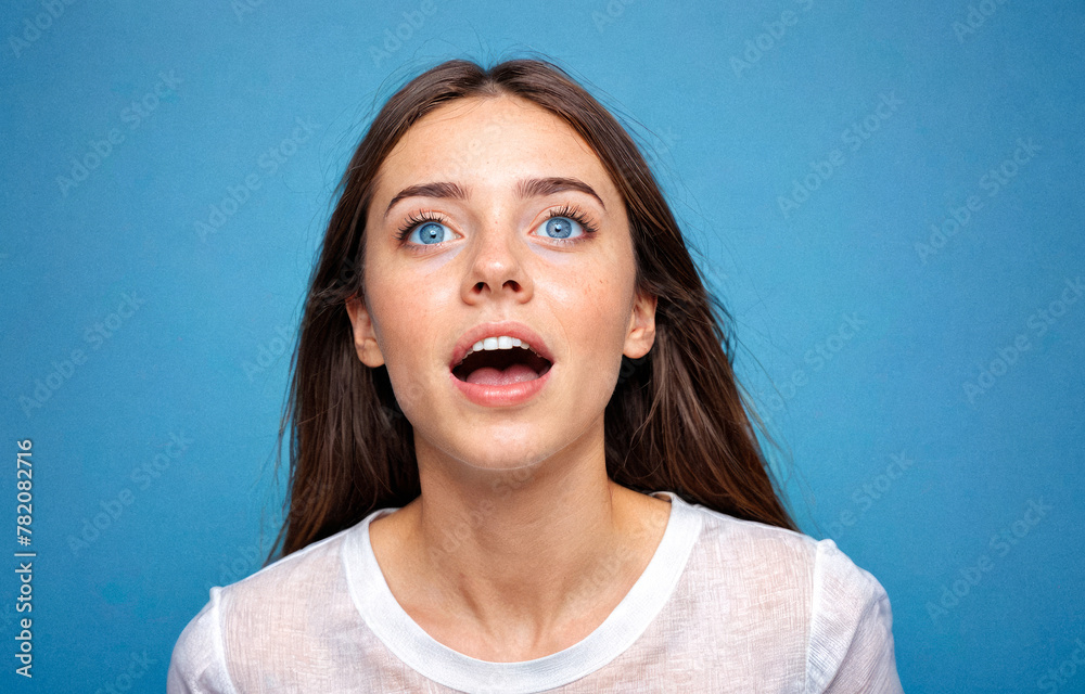 Young woman, pleasantly surprised, showcases big eyes, open mouth. Captured against vibrant blue background, expressive facial features highlighted. Wearing casual white top Young woman, pleasantly surprised, showcases big eyes, open mouth. Captured against vibrant blue background, expressive facial features highlighted. Wearing casual white top