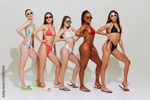 Young slim girls with different skin tones posing holding hand on hip in line swimsuit and against white studio background. Concept of summer holidays, recreation, vacation, luxury resort. Ad