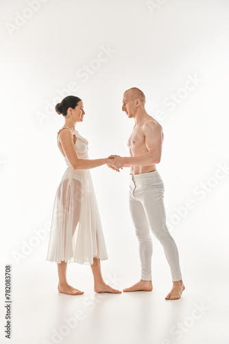 A young shirtless man and a woman in a white dress unite in a graceful dance, intertwining in acrobatic elements against a white backdrop.