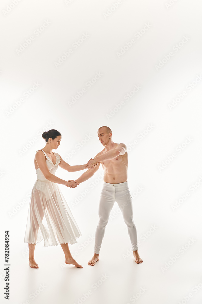 A young shirtless man and a young woman in a white dress dance together, executing acrobatic elements in a studio setting on a white background.