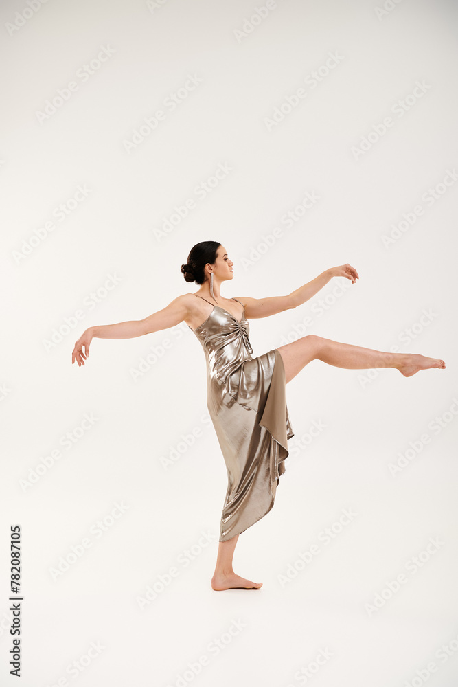 A young woman exudes grace and elegance as she dances in a long, shiny silver dress in a studio setting against a white backdrop.